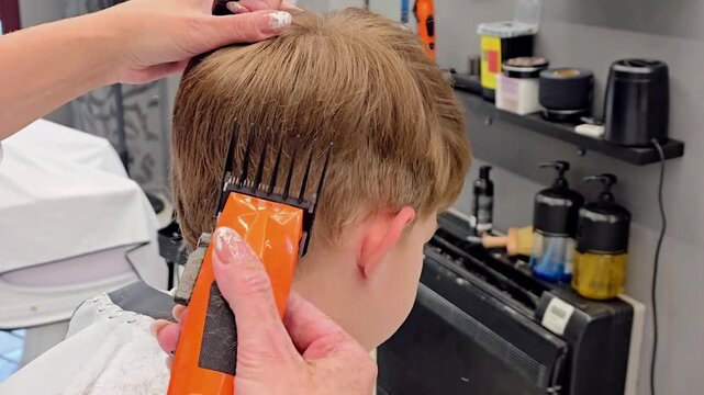 A young boy receives a haircut at a barbershop as stylist uses an orange electric clipper to carefully trim the hair around his head, showing professional grooming and salon work in progress