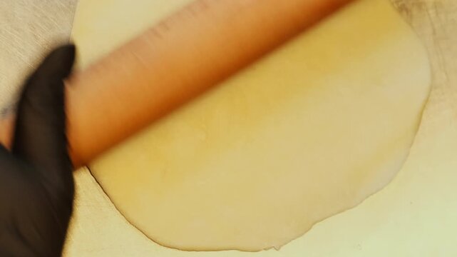 Baker rolls pale pastry dough across floured surface with wooden pin. Repeated strokes flatten soft sheet evenly preparing wrapper for filling