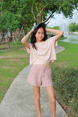Fototapeta premium A young Asian girl wearing a casual pink top, shorts, and a cap posing on a pathway in a park.