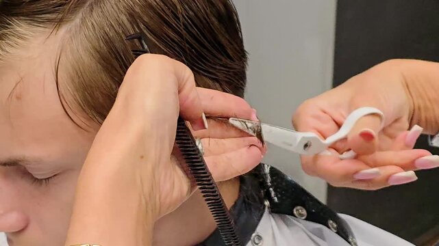 Barber cutting a teenager's hair in a barbershop, using scissors and comb during a professional haircut, with salon tools and equipment clearly visible in the background in the salon setting