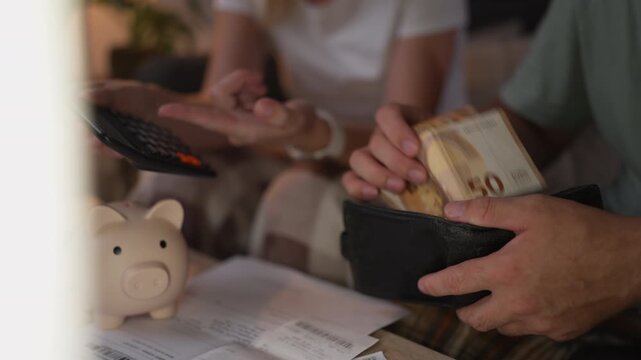 Close-up of a couple managing their monthly budget at home, a woman using a calculator while a man takes fifty euro banknotes from his wallet to pay bills or save money in a piggy bank
