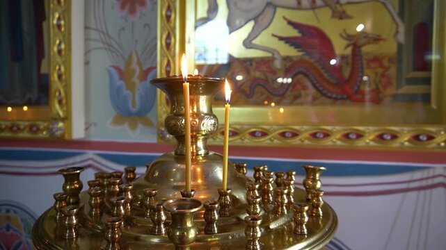 Lit candles on brass church candle stand. Orthodox temple interior with gilded icon frame and wall painting, two burning thin candles on metal holder, worship and prayer concept, no people.