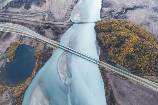 Aerial view of the Knik River with a highway bridge crossing the glacial blue water, surrounded by autumn foliage and rugged terrain in Knik River, Alaska, United States.