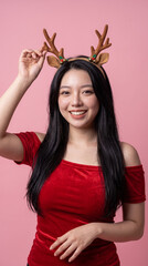 Happy asian woman wearing reindeer antlers headband posing on pink background © iz_zi