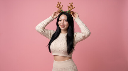 Happy asian woman wearing reindeer antlers headband posing on pink background © iz_zi