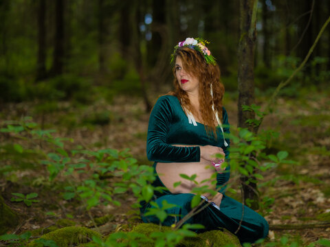 Expectant Mother Holding Twin Baby Shoes on Pregnant Belly in Woodland. Beautiful pregnant woman with long red hair sitting front-facing on a moss-covered stump in a forest. Her hands rest gently over