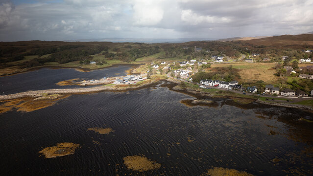 Aerial view of the coastal village of Arisaig with its harbor, scattered houses, and green hills under a cloudy sky in Arisaig, Scotland, United Kingdom.