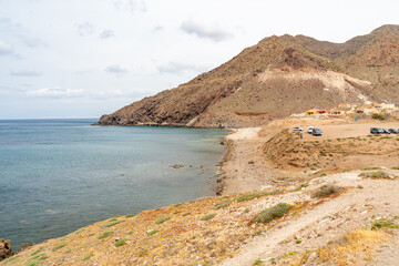 Arid landscape with rugged hills meeting a calm blue sea, a secluded beach, and a small settlement with parked vehicles, conveying themes of nature, travel, and adventure © Osvaldo Mussi