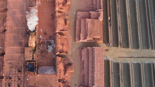 Top-down drone view of a brick field in Bangladesh with raw drying bricks, stacks of fired bricks and smoke rising from a traditional kiln.