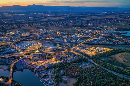 Aerial view of the city of Mulhouse at dusk, featuring illuminated streets, industrial areas, and a nearby lake, set against the backdrop of distant mountains in Mulhouse, Grand Est, France.