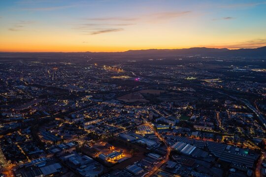 Aerial view of the city of Mulhouse at twilight, with glowing urban lights, residential areas, and distant mountains under a sunset sky in Mulhouse, Grand Est, France.