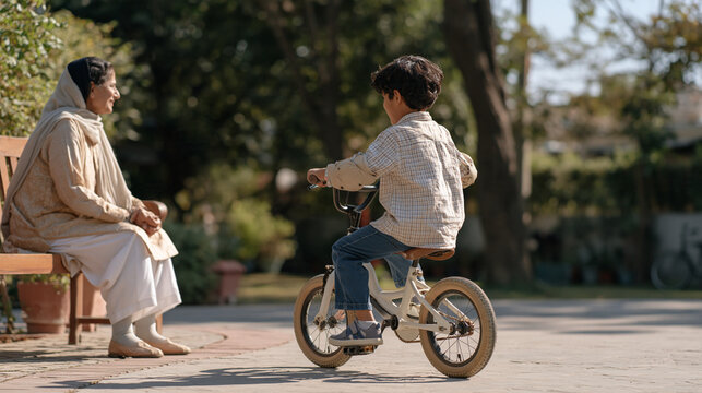 Pakistani grandmother sitting on a garden bench watching her grandson practice slow pedaling in a circular driveway, protective padding visible on elbows and knees,