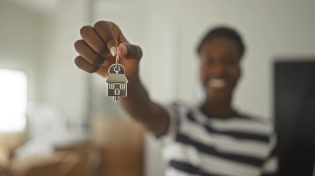 Young african american man holding a house key keychain between fingers, hand extended toward camera in residential building; new home pride.