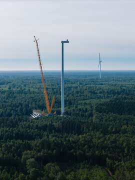 Aerial view of a wind turbine under construction with a large yellow crane in a forest clearing in Ventspils, Ventspils Municipality, Latvia.