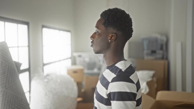 Man in striped shirt stands looking at packed moving boxes and bubblewrap, face in profile in building; quiet uncertainty.