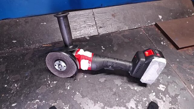 Angle grinder stays on gritty workshop floor next to steel offcuts. Battery powered grinder remains idle with taped body and mounted wheel, ready for metalworking tasks in practical workspace.