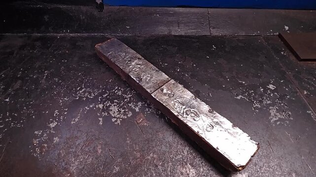 Forged metal bar lies on dark workshop surface in closeup. This metal piece catches light along its textured face, suggesting careful handling and moody industrial atmosphere with restrained energy.