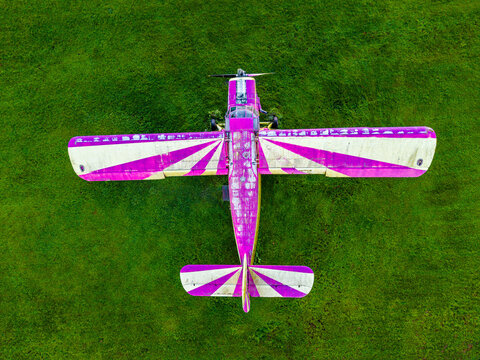 Aerial view of a purple and white striped propeller plane parked on a vibrant green grass field in Latvia.