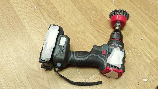 Cordless drill lies on wooden floor, equipped with brush attachment and taped battery in tool setup. Tool appears prepared for cleaning duty, with compact handheld power unit and visible strap.