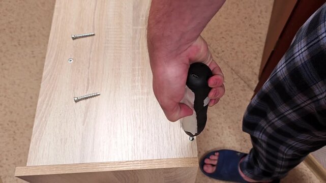 Furniture assembly starts with screwdriver and wood panel, as hands drive screws into drilled holes. This focused home task shows practical setup work on light colored board with loose fasteners.