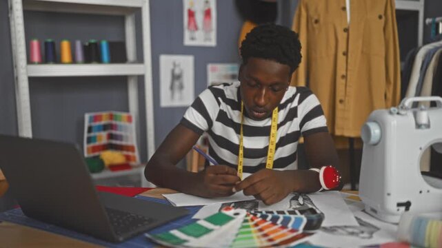 Man sketching on paper with laptop open and measuring tape around neck, hands holding pencil in studio; creativity concentration.