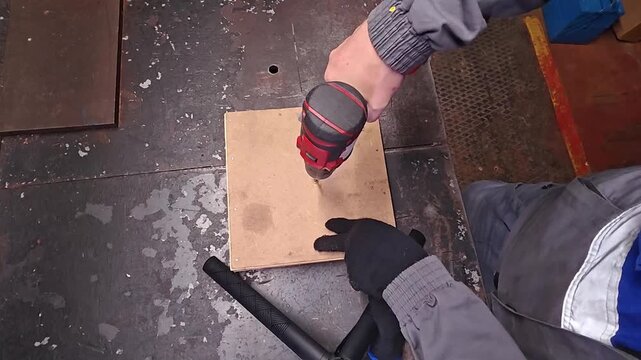 Wood drilling in workshop as gloved hands guide power drill into board. Worker steadies wood on workbench, creating clean hole during careful drilling practice with focused effort.