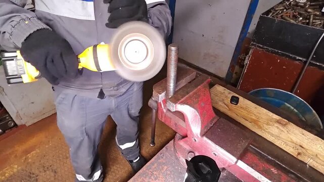 Gloved worker operates yellow angle grinder on clamped metal stock inside workshop. Metal grinding happens beside red vise and wooden block, creating practical repair and fabrication task.