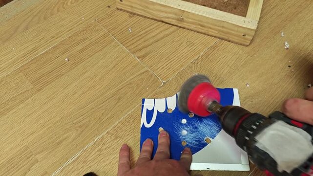 In home workshop, hands guide spinning drill brush over blue surface panel with steady control. Tool user works carefully on coated piece, showing concentrated DIY detail on wood flooring.