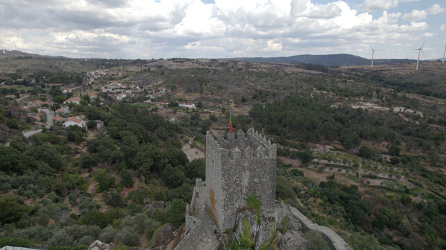 Aerial view of the medieval stone castle tower of Sortelha, surrounded by lush green hills, olive groves, and distant wind turbines in the rural landscape of Sortelha, Distrito da Guarda, Portugal.