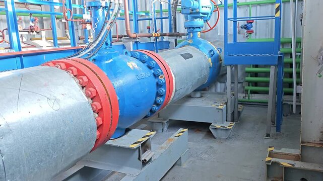 Blue industrial pipe assembly fills plant room interior. Large bolted flanges, valves and green guard rails frame machinery, presenting orderly equipment interior with clean painted surfaces.