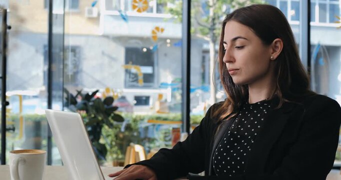 Concentrated female freelancer uses computer at cafe table near large window. Elegant young woman pauses typing occasionally to look away thoughtfully before continuing remote project.
