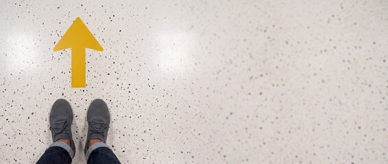 Fototapeta premium High angle POV of person in grey sneakers standing in front of a yellow directional arrow on a terrazzo floor, symbolizing progress, future goals, and moving forward with copy space.