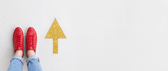 Fototapeta premium Top view of a person wearing red sneakers standing next to a golden arrow pointing forward on a neutral background with large negative space on the right for text, representing choice and direction