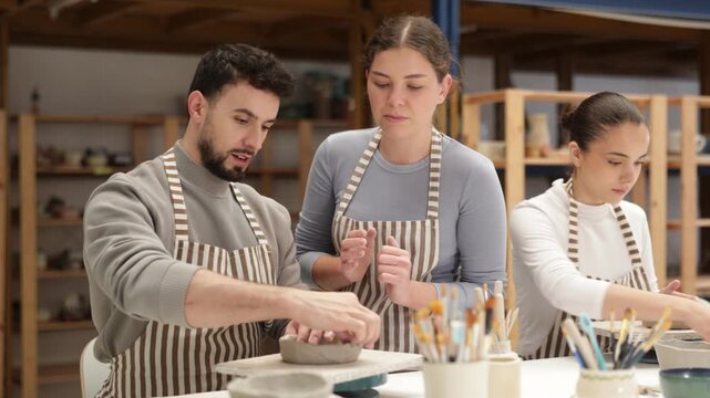 Man craftsperson in striped aprons busy molding clay products in lesson with girl teacher. Amateur sculpting plate of raw clay under supervision of teacher 