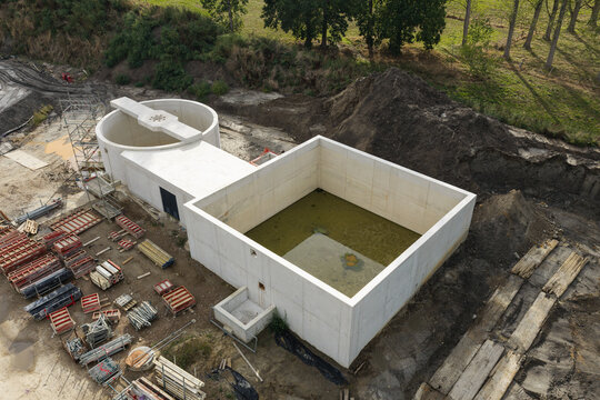 Aerial view of a wastewater treatment plant construction site with a large concrete settling tank and circular basin, scaffolding, and industrial equipment in Diepenbeek, Flanders, Belgium.