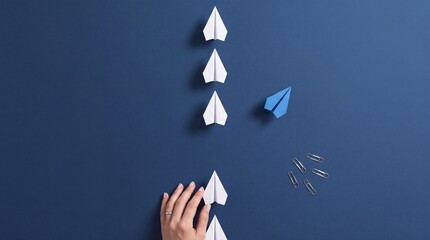 Fototapeta premium Top Down View of a Hand Placing White Paper Planes in a Row with One Blue Paper Plane Changing Direction on a Dark Blue Background, Concept of Leadership, Uniqueness, and Business Innovation