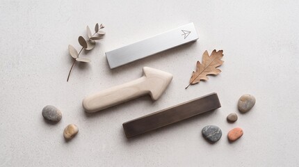 Fototapeta premium Overhead flat lay of minimalist furniture hardware including metal cabinet pulls, a wooden arrow, smooth river stones, and dried autumn leaves on a light textured background.