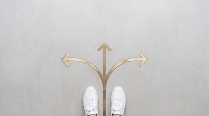Fototapeta premium Top view of a person wearing white sneakers standing at a three-way arrow crossroads on a concrete floor, representing decision making, career path options, and choosing the right direction.