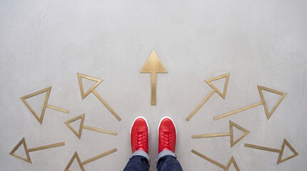 Fototapeta premium High angle top down view of a person wearing red sneakers standing on a concrete floor surrounded by multiple golden arrows pointing in different directions representing choice and career path.