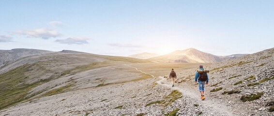 Fototapeta premium Rear view of two adventurous male hikers trekking along a winding stone path in a vast, barren mountain range under a clear sky with sunlight, ideal for travel, exploration, and outdoor lifestyle.