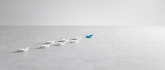 Fototapeta premium A blue origami paper boat leading a group of white paper boats on a gray textured background, representing leadership, business success, unique vision, and individuality in a minimalist wide