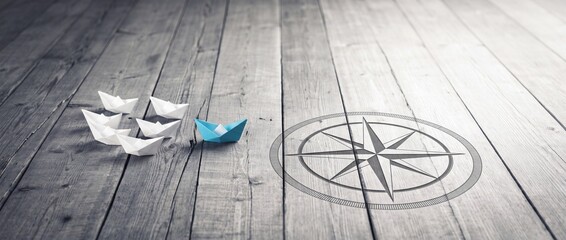 Fototapeta premium Blue Origami Paper Boat Leading a Fleet of White Boats Across a Wooden Surface Toward a Compass Symbol, Representing Leadership, Strategic Business Direction, and Vision in a Wide Panorama