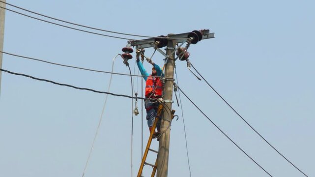 An electrician working on a newly installed utility pole is installing cables on insulators and pulling them with a hoist.
