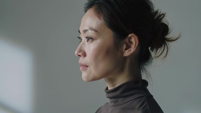 Turning Asian woman in studio wearing dark turtleneck, responding to left window light, copy space