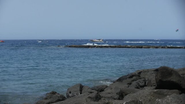 Speedboat racing across deep blue ocean water beyond a rocky breakwater, leaving a white wake behind. Wide coastal view with shoreline rocks, small boats, and parasailing