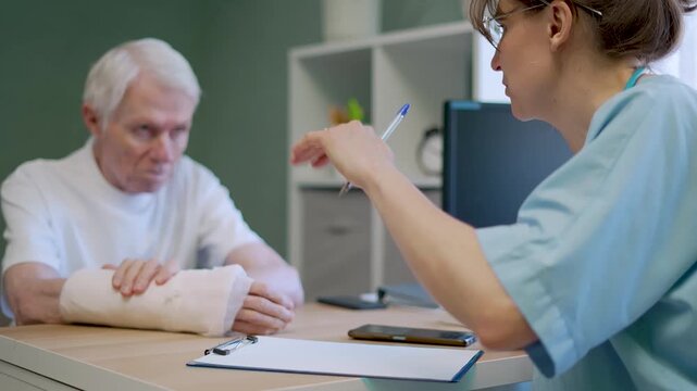 Anxious senior man discusses treatment with female doctor in clinic. Elderly patient worries about cast while medical professional explains care plan for injured arm.
