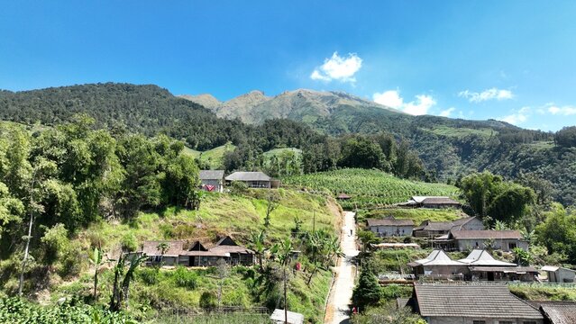 Aerial view of KEMA MERBABU Glamour Camping nestled on the slopes of Mount Merbabu with traditional houses, terraced fields, and blue sky in Kecamatan Ampel, Jawa Tengah, Indonesia.