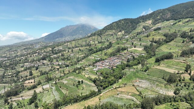 Aerial view of KEMA MERBABU Glamour Camping nestled among lush green terraced fields and a small village on the slopes of Mount Merbabu in Kecamatan Ampel, Jawa Tengah, Indonesia.