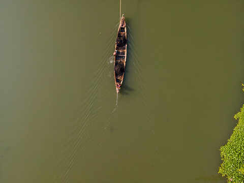 Aerial view of a small wooden boat with a person navigating a calm river, with lush green vegetation along the bank in Patikraja, Central Java, Indonesia.