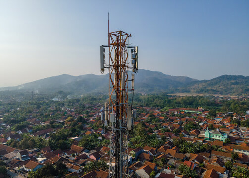 Aerial view of a telecommunications tower rising above a residential neighborhood with red-tiled roofs and greenery against a backdrop of mountains in Patikraja, Central Java, Indonesia.